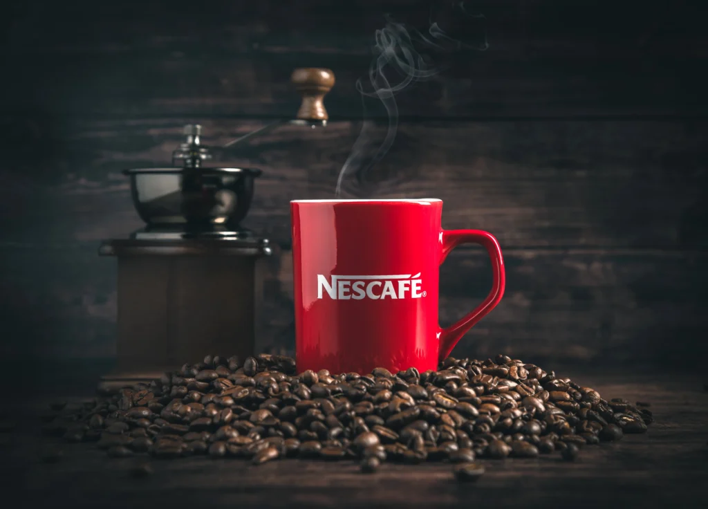 A steaming cup of Nescafe coffee sits on a saucer, surrounded by coffee beans and a vintage coffee grinder in the background